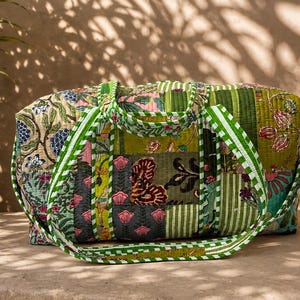 May include: A quilted duffel bag featuring a patchwork design with various floral and striped patterns in shades of green, pink, and cream. The bag includes two green and white striped fabric handles and a matching striped shoulder strap.