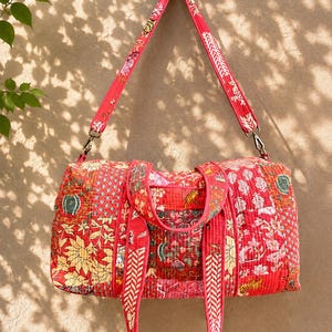 May include: A quilted duffel bag featuring a vibrant red patchwork design with various floral and animal patterns. The bag includes two short carrying handles and a long, adjustable shoulder strap, both matching the red patterned fabric.