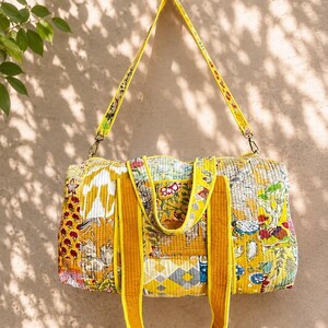 May include: A quilted duffel bag featuring a patchwork design in shades of yellow, white, and multi-coloured floral patterns. The bag includes two solid yellow top handles and a detachable, adjustable yellow shoulder strap with metal hardware.