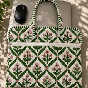 Op de afbeelding: Een gewatteerde laptophoes met een witte achtergrond, een groen ruitpatroon en roze bloemmotieven. De hoes is voorzien van twee handvatten, een witte ritssluiting aan de voorzijde en een bijpassende groen-witte gestreepte rand.