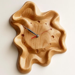 Könnte beinhalten: Eine Wanduhr aus hellem Holz mit einer unregelmäßigen, organischen, wellenförmigen Form. Das Zifferblatt hat eine glatte, geschnitzte Oberfläche mit kleinen kreisförmigen Vertiefungen als Stundenmarkierungen, schwarzen Zeigern und einem roten Sekundenzeiger.