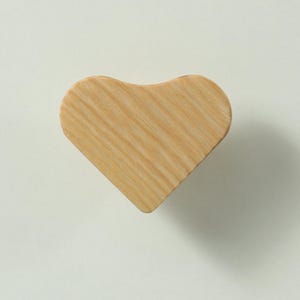 May include: A heart-shaped wooden knob with a light tan colour and a visible natural wood grain pattern. The object has a smooth, flat surface and a rounded top edge, showcasing the organic texture and light brown tones of the wood material.
