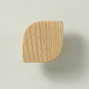 May include: A light brown wooden cabinet knob with a smooth, curved, leaf-like shape. The natural wood grain is visible across the surface, showing vertical lines and patterns. The knob has a flat front face with rounded edges on one side.
