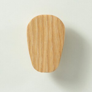 May include: A light brown wooden wall hook with a smooth, rounded top and a tapered base. The natural wood grain is visible across the surface, showing vertical lines and a light tan colour. The object has a flat front face and a solid construction.