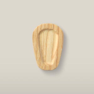 Puede incluir: Una bandeja de madera de color claro con una forma suave, redondeada y similar a un escudo. La superficie presenta un patrón de veta de madera natural visible que corre verticalmente, con un área central poco profunda y hundida.