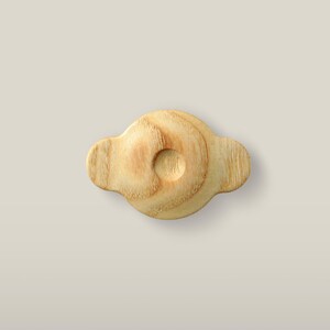 May include: A light-colored wooden knob with a smooth, rounded central section and two protruding side tabs. The natural wood grain is visible across the surface, and a small, circular indentation is centered on the top of the knob.