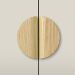 May include: Two semi-circular light wood cabinet pulls mounted on a light beige surface. The handles are positioned vertically, facing each other to form a complete circle. The natural wood grain is visible, running vertically along the surface.