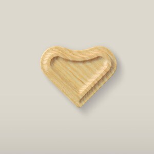 Può includere: Un piattino in legno a forma di cuore di colore marrone chiaro con venature naturali del legno visibili. L'oggetto presenta una superficie interna liscia e scavata con un bordo rialzato, che mette in risalto la texture organica.