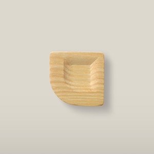 Pode incluir: Um pequeno prato de madeira de cor clara com um padrão de grão natural e suave. O objeto apresenta uma reentrância central de forma quadrada com cantos arredondados e uma borda externa plana e curva, exibindo a textura orgânica da madeira.