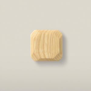 Pode incluir: Um puxador quadrado de madeira com bordas chanfradas e um acabamento de veios de madeira natural em tom castanho claro. O objeto apresenta uma superfície superior plana e lados inclinados que se estreitam para a base, mostrando a textura.