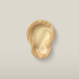 May include: A small, light-coloured wooden dish carved into the shape of a human ear. The object features a smooth, polished surface with visible natural wood grain patterns running throughout the light tan and beige material of the carved form.