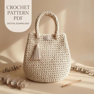 Könnte beinhalten: Eine beige gehäkelte Handtasche mit geflochtenem Griff und passendem Quastenanhänger. Die Tasche hat ein kleines rechteckiges Etikett auf der Vorderseite. Der Text oben links lautet: CROCHET PATTERN PDF DIGITAL DOWNLOAD. Eine Holzhäkelnadel liegt dabei.