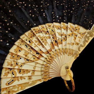 Può includere: Un ventaglio pieghevole con una copertura in pizzo nero decorata con piccole paillettes nere. Le stecche sono realizzate in materiale bianco sporco con intricati motivi in rilievo floreali e a volute color oro. Un anello in metallo dorato è fissato alla base.