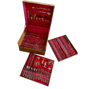 May include: A vintage wooden cutlery set with a red velvet interior. The set includes various gold-colored utensils, such as knives, forks, and spoons, neatly arranged in a wooden box and two trays. The set also includes serving pieces.
