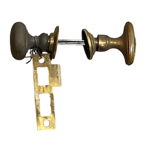 Puede incluir: Juego de manijas de puerta de latón antiguo con un pomo ovalado marrón oscuro, una placa redonda de latón y una placa de cierre rectangular de latón. El juego incluye una varilla de conexión y está aislado sobre un fondo blanco.