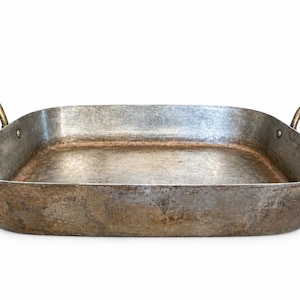 May include: A rectangular metal roasting pan with a weathered, tarnished silver and brown patina. The pan features two curved metal handles attached to the sides with rivets, and the interior base shows signs of heavy use and discoloration.