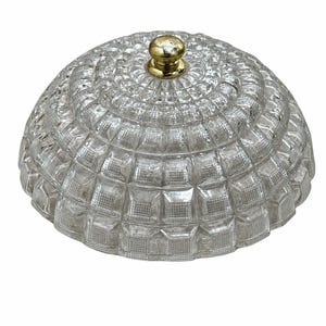 May include: A clear, dome-shaped glass lid featuring a textured, grid-like pattern of raised squares across its surface. The lid is topped with a small, round, metallic gold-colored knob handle centered at the very peak of the dome.