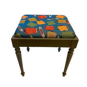 May include: A small, green wooden stool with turned legs. The seat is upholstered in a blue fabric with a pattern of colorful spools of thread.