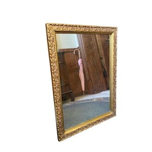 May include: A rectangular gold-framed mirror with ornate floral detailing. The mirror reflects a room with wooden furniture and a pink umbrella. The frame has a warm, antique gold finish, adding a touch of elegance.