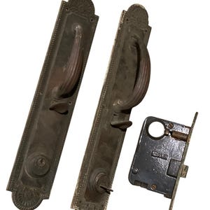 May include: Two antique brass door handles with ornate details and a matching brass door lock. The handles have a curved design and are attached to rectangular plates with decorative borders. The lock is a separate piece with a keyhole and a latch.