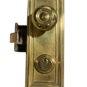 Könnte beinhalten: Ein antikes Türschloss aus Messing mit rundem Knauf und Schlüsselloch. Die rechteckige Platte hat eine strukturierte Oberfläche und einen dekorativen Rahmen. Der Schließmechanismus ist seitlich sichtbar.