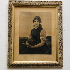 May include: A framed black and white photograph of a young woman wearing a simple dress and a headscarf. She is sitting in a field and looking directly at the viewer.