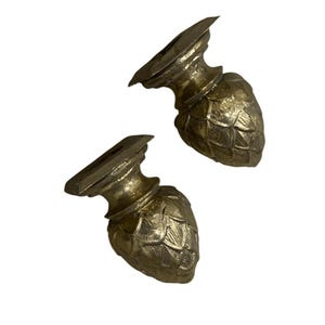 May include: Two gold-colored, decorative furniture feet. Each foot has a detailed, artichoke-like base and a rectangular top with a recessed area. The feet are made of a metallic material and are designed to be attached to furniture.