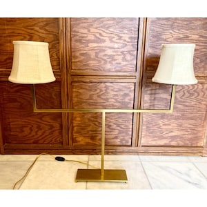 May include: A brass-toned table lamp with two cream-colored lampshades. The lamp has a rectangular base and a horizontal bar supporting the shades. The cord and plug are visible. The background is a wooden wall.