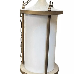 May include: A brass hanging light fixture with a white frosted glass cylinder shade. The fixture has a chain and a decorative top.