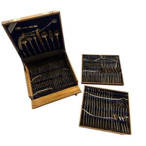 May include: A wooden silverware chest with a blue velvet interior, open to reveal a complete set of silver utensils. Additional trays of silverware are displayed alongside the chest. The set includes forks, knives, spoons, and serving pieces.