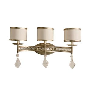 May include: A three-light wall sconce with a silver finish. The sconce features three cream-colored lampshades with gold trim. A row of clear crystals decorates the center, and clear crystal pendants hang below the sconce.