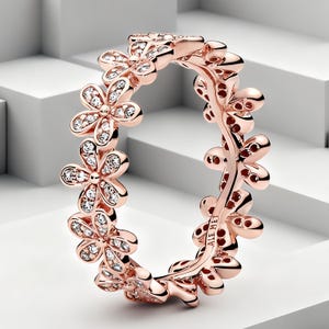 Puede incluir: Un anillo en tono oro rosa que presenta una banda continua de formas de flores interconectadas. Cada flor está detallada con pequeñas piedras claras y brillantes incrustadas en los pétalos. La banda interior tiene grabado ALE MET.