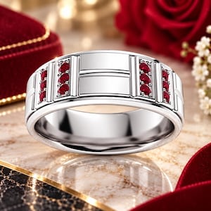 Op de afbeelding: Een gepolijste zilverkleurige ring met aan weerszijden twee verticale kanalen, elk bezet met drie kleine ronde rode robijnen. Het midden van de ring toont een glad, plat rechthoekig paneel met een horizontale lijndetail.