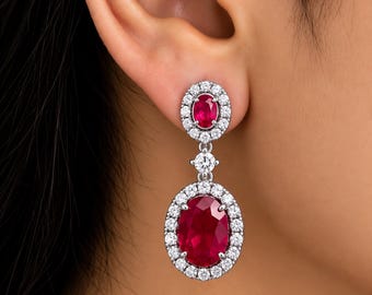 May include: A drop earring featuring a small oval ruby surrounded by a halo of white diamonds, connected by a single round diamond to a larger oval ruby drop, also encircled by a halo of white diamonds, all set in polished silver-toned metal.