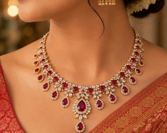 May include: A gold-toned necklace and matching jhumka earring set featuring teardrop-shaped red stones surrounded by clear white crystals. The necklace has a central pendant with a dangling teardrop stone and a symmetrical floral-inspired design.