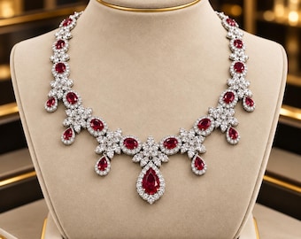 Puede incluir: Un collar de tono plateado con múltiples rubíes rojos ovalados y en forma de pera, rodeados de pequeñas piedras claras y brillantes. El diseño incluye un colgante central en forma de pera con racimos florales simétricos de piedras.
