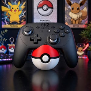 Può includere: Un controller Nintendo Switch Pro nero appoggiato su un supporto sferico rosso e bianco a tema Pokeball. Il controller presenta pulsanti neri standard, due levette analogiche, un pad direzionale e il logo Nintendo Switch stampato in bianco.