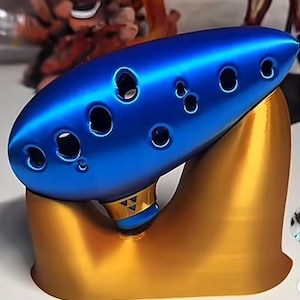 Peut inclure: Un ocarina bleu métallique doté de plusieurs trous pour les doigts, posé sur un support doré lisse et incurvé. L'instrument présente une petite bande dorée près de la base avec un symbole triangulaire gravé, affichant une finition élégante.