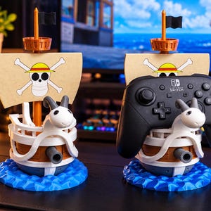 Peut inclure: Deux modèles de navires imprimés en 3D avec une figure de proue en forme de chèvre blanche, une coque marron et une voile beige ornée d'un crâne avec un chapeau de paille. L'un des modèles porte une manette Nintendo Switch noire sur son pont.