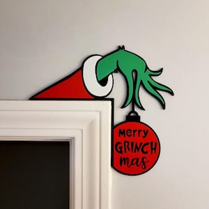 Peut inclure: Une décoration d'angle de porte représentant une main verte sortant d'un bonnet rouge et blanc. La main tient une boule rouge avec le texte noir merry GRINCH mas. Le design est plat avec des contours noirs et des couleurs vives.