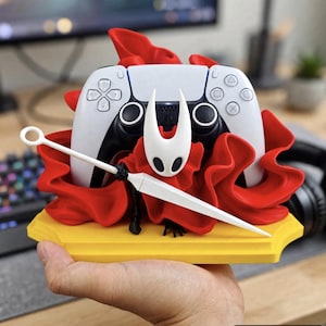 Puede incluir: Un soporte para mando de videojuegos en blanco y negro que presenta una máscara de personaje blanca con un arma de aguja puntiaguda y elementos de tela roja fluida, todo montado sobre una base hexagonal amarilla sólida.