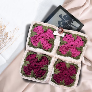Op de afbeelding: Een vierkante gehaakte tablethoes met vier bloemige granny squares in tinten magenta en diep bordeaux met olijfgroene bladeren, omrand met crèmekleurig garen. Een donkere houten knoop sluit de flap en er is een klein wit label bevestigd.