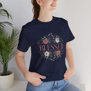 May include: A navy blue short-sleeve t-shirt featuring a central graphic design. The design consists of a circular arrangement of botanical elements, including pink and white roses and green leaves, surrounding the word BLESSED in pink text.