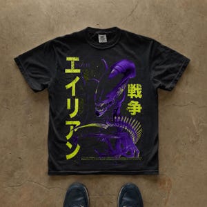 Puede incluir: Una camiseta negra de manga corta con un gran gráfico morado de una criatura xenomorfa. El diseño incluye caracteres japoneses de color amarillo brillante dispuestos verticalmente a los lados, con un pequeño texto blanco en la parte inferior central.