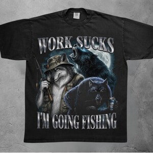以下が含まれることがあります： 3匹のオオカミのグラフィックが描かれた黒いTシャツ。1匹は釣り用の帽子をかぶって釣り竿を持ち、もう1匹は月に吠え、3匹目は缶を持っています。WORK SUCKSとI'M GOING FISHINGという文字がメタリックシルバーのフォントでプリントされています。