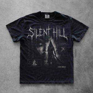May include: A black short-sleeved t-shirt featuring a white graphic print of the Silent Hill logo at the top. The design includes a central illustration of a pyramid-headed figure, a faint face in the background, and small technical grid details.