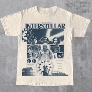 Op de afbeelding: Een crèmekleurig t-shirt met korte mouwen en een donkerblauwe grafische print geïnspireerd op de film Interstellar. Het ontwerp bevat een collage van filmscènes, een grote klok, de filmtitel en de namen van de castleden.