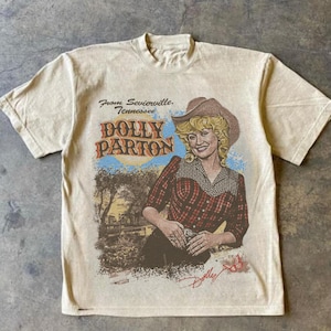 May include: A beige short-sleeve t-shirt featuring a graphic print of Dolly Parton wearing a cowboy hat and a red plaid shirt. The text reads From Sevierville, Tennessee, Dolly Parton, and includes a small butterfly illustration at the bottom.