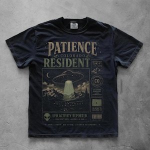 Könnte beinhalten: Ein dunkelgraues Kurzarm-T-Shirt mit einem Grafikdruck und dem Text PATIENCE COLORADO RESIDENT. Das Design zeigt eine fliegende Untertasse über Bergen, einen Alien-Kopf sowie verschiedene Informationsfelder mit Koordinaten und Höhenangaben.