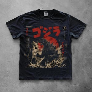 Könnte beinhalten: Ein schwarzes Kurzarm-T-Shirt mit einer Grafik von Godzilla, der vor einer großen roten Sonne aus dem Wasser auftaucht. Das Design enthält japanischen Text, die Silhouette eines Hubschraubers und eine Stadtsilhouette in einem abgenutzten Druck.